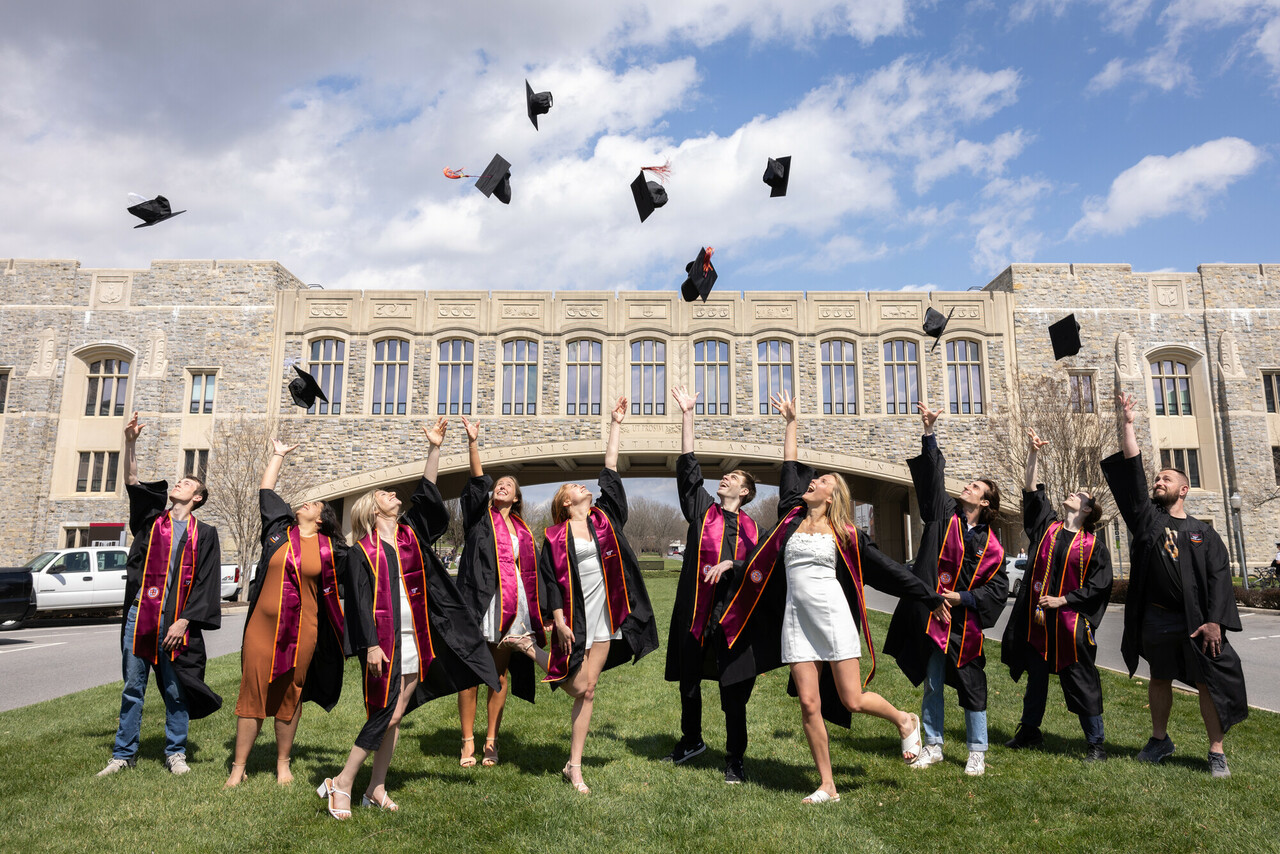 Group of various students, standing on grass, wearing graduation attire, throwing their caps in the air in front of Torgersen bridge