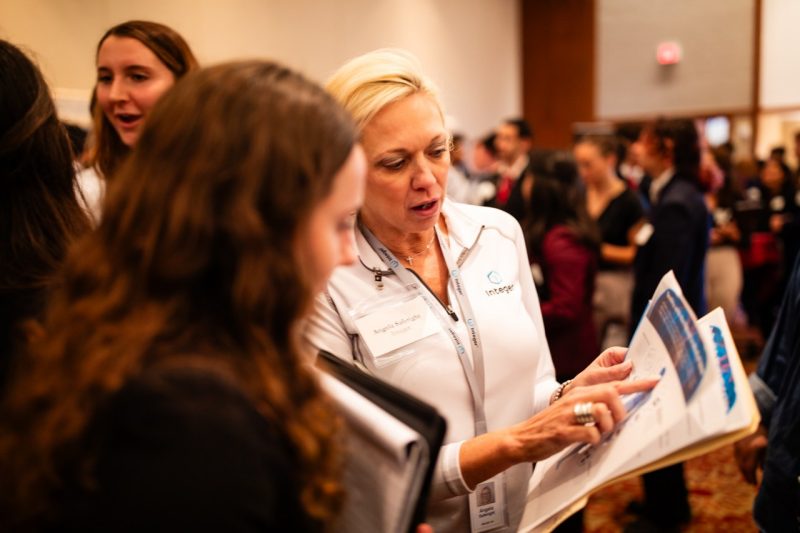 Femal student with dark curly hair, holding a padfolio, listens while a blonde woman, wearing a white shirt points to a piece of paper