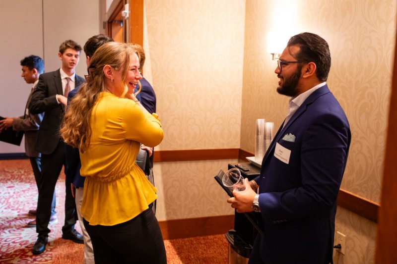 Young woman with long blonde hair, wearing a yellow shirt talks with a beareded gentleman wearing glasses and a dark suit.