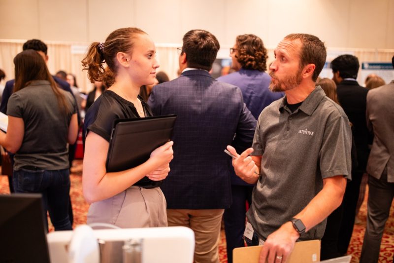Young woman with dark hair pulled into a ponytail, wearing a blue short sleeved shirt talks with a bearded man wearing a gray polo.