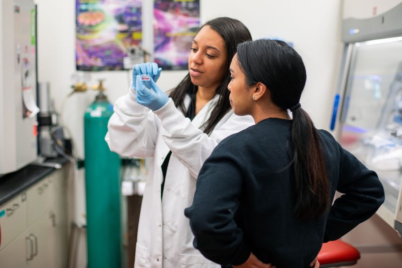 Portrait of Michelle standing in a lab, showing another student a piece of equipment.