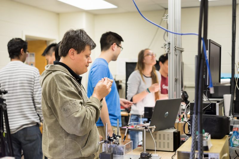 A group of students stands in a lab, among bars and wires and other engineering materials