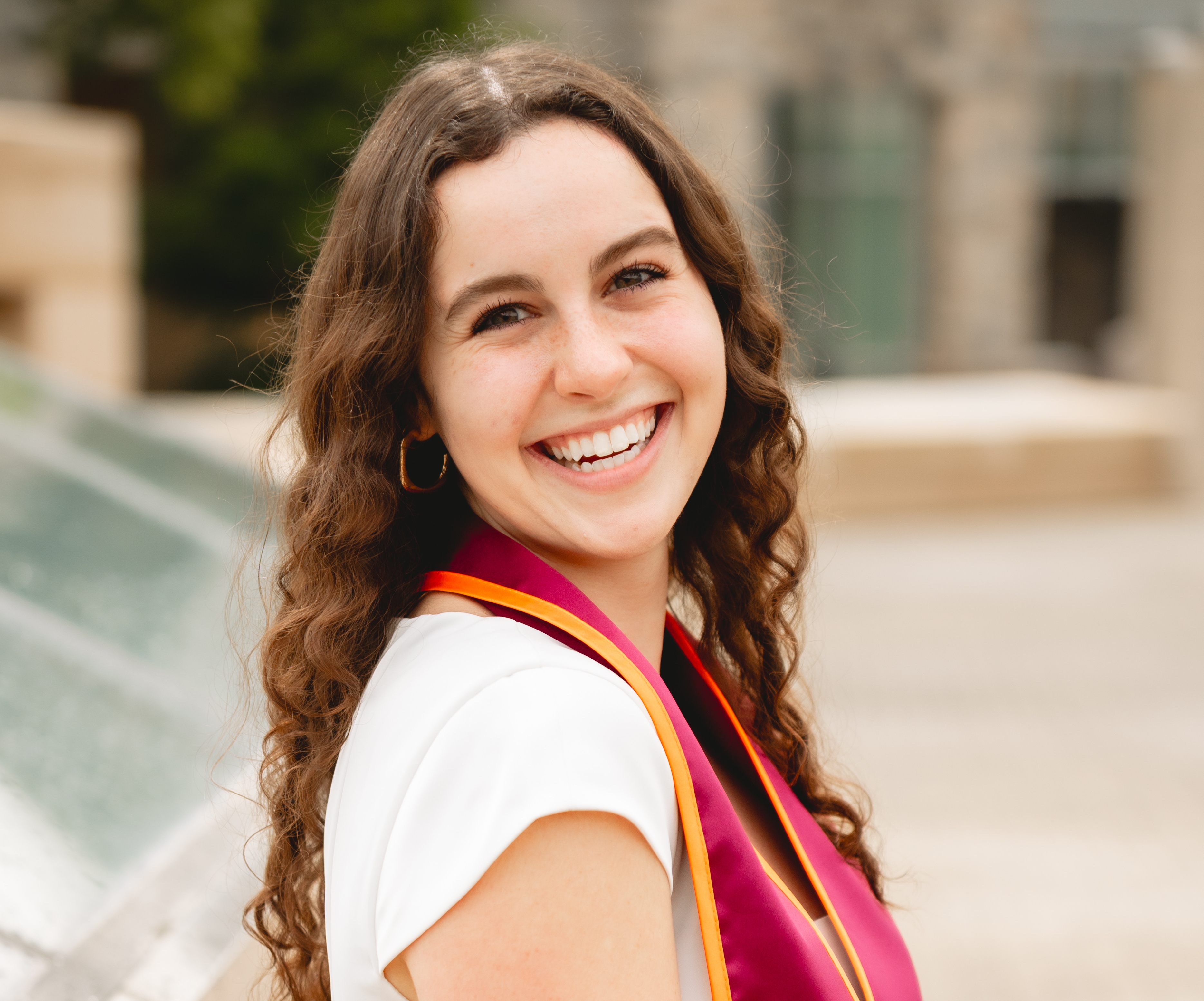 Young woman with long, dark curly hair wearing a white dress with her graduation stole