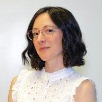 Woman with shoulder length brown wavy hair, wearing glasses, and a white, sleeveless shirt.