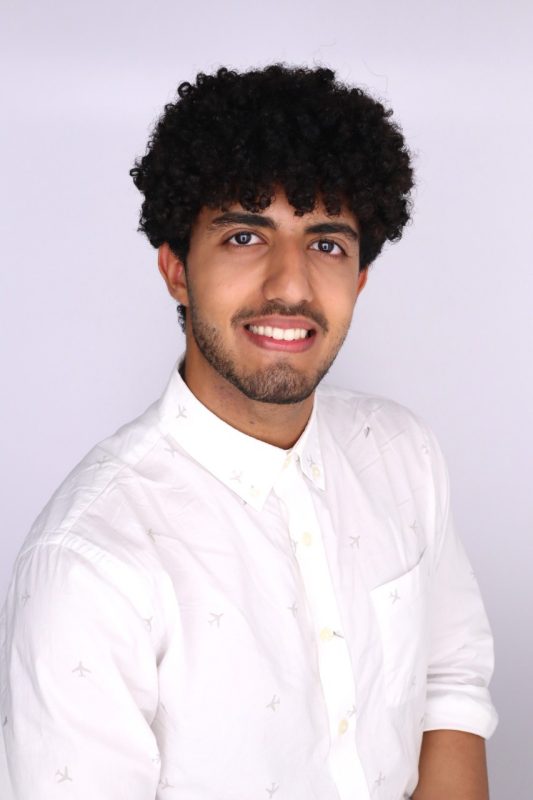 Young man with dark curly hair, a light beard, wearing a white button down collared shirt.