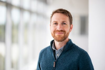 Man with reddish hair and beard, wearing a blue 1/4 zip sweater in front of a bank of windows