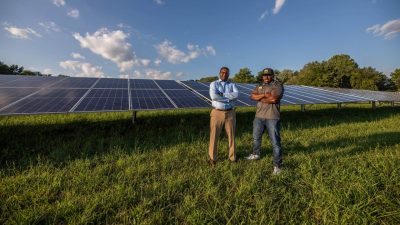 Two men pose in front of solar panels in a field.