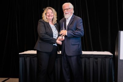 A woman with blonde hair, wearing a dark suit presents a crystal award to a man with a white beard and hair, wearing a dark suit. 