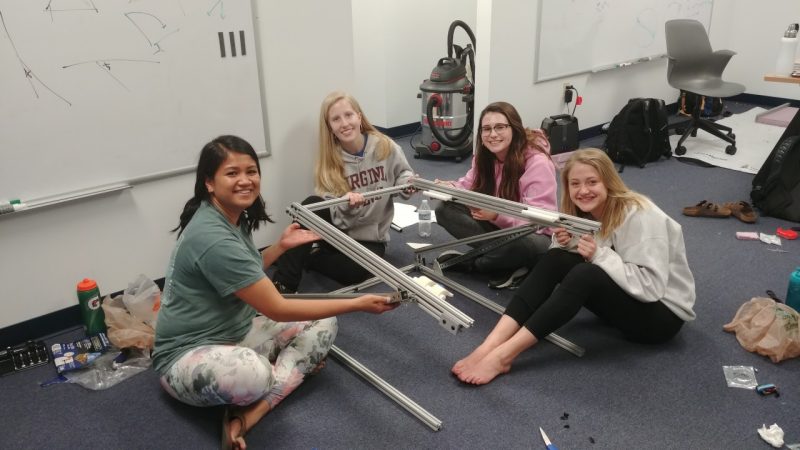 Four girls are sitting on the floor smiling as they hold up a metal device.