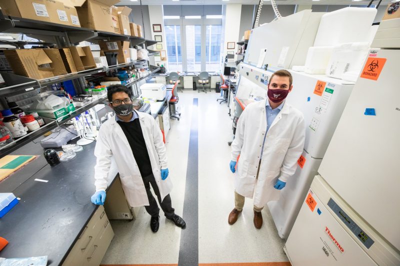 Doctoral students Graybill and Jana stand in a research lab, with masks and lab coats on.