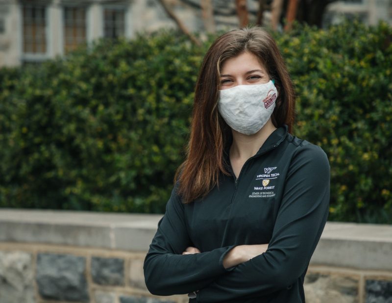 Nicole Stark, doctoral student in biomedical engineering and mechanics, stands in front of green bushes on Virginia Tech campus, wearing a black VT pullover and a mask.